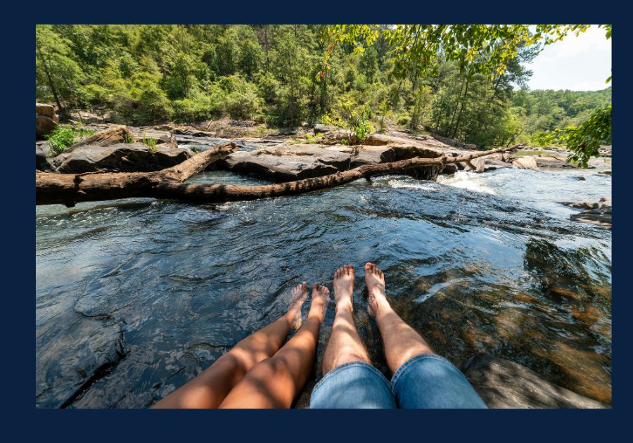 Two people sitting with their feet in a clear East Tennessee creek surrounded by rocks, trees, and flowing water.